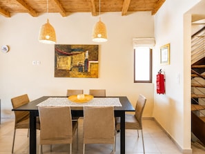 Uma sala de jantar com mesa, cadeiras, luminárias pendentes, um quadro, um extintor de incêndio e uma escada.