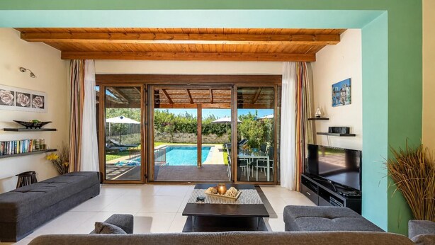 A living room with a pool, sliding glass doors, a flat-screen TV, and a sofa. - Villa Mandarin - Two Bedroom Villa, Sleeps 4 (Platanias)