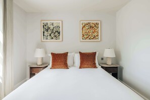 Habitación de hotel con una cama grande, dos mesitas de noche, dos lámparas fijadas a la pared y dos obras de arte enmarcadas sobre la cama.