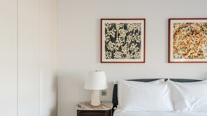 Een slaapkamer met een bed, een nachtkastje met een lamp, en twee ingelijste bloemstillevens aan de muur.