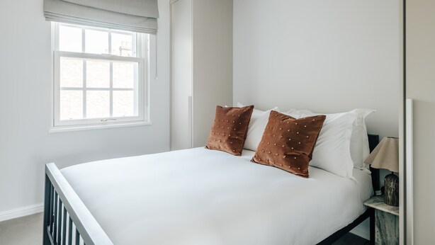 Et soveværelse med en seng, to puder, et natbord, en lampe og et vindue med persienner.