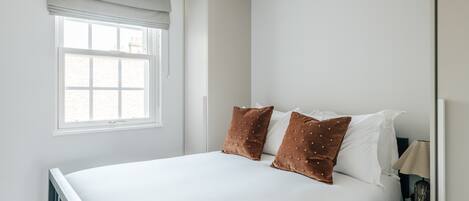 Ein Schlafzimmer mit einem Bett, zwei Kissen, einem Nachttisch, einer Lampe und einem Fenster mit Jalousien.