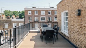 En balkon med et bord og stole, en murstensvæg og en tilstødende bygning med mange vinduer.