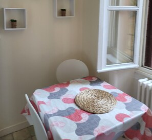 A small room with a table covered in a patterned tablecloth, a woven basket, and a white chair. There are two wall-mounted shelves with plants. - Quiet apartment near amenities, located on a cul-de-sac (Aurillac)
