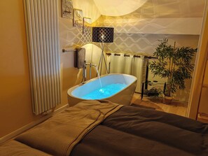 A modern bathroom with a freestanding bathtub, a towel rack, a potted plant, and a wall-mounted light fixture.