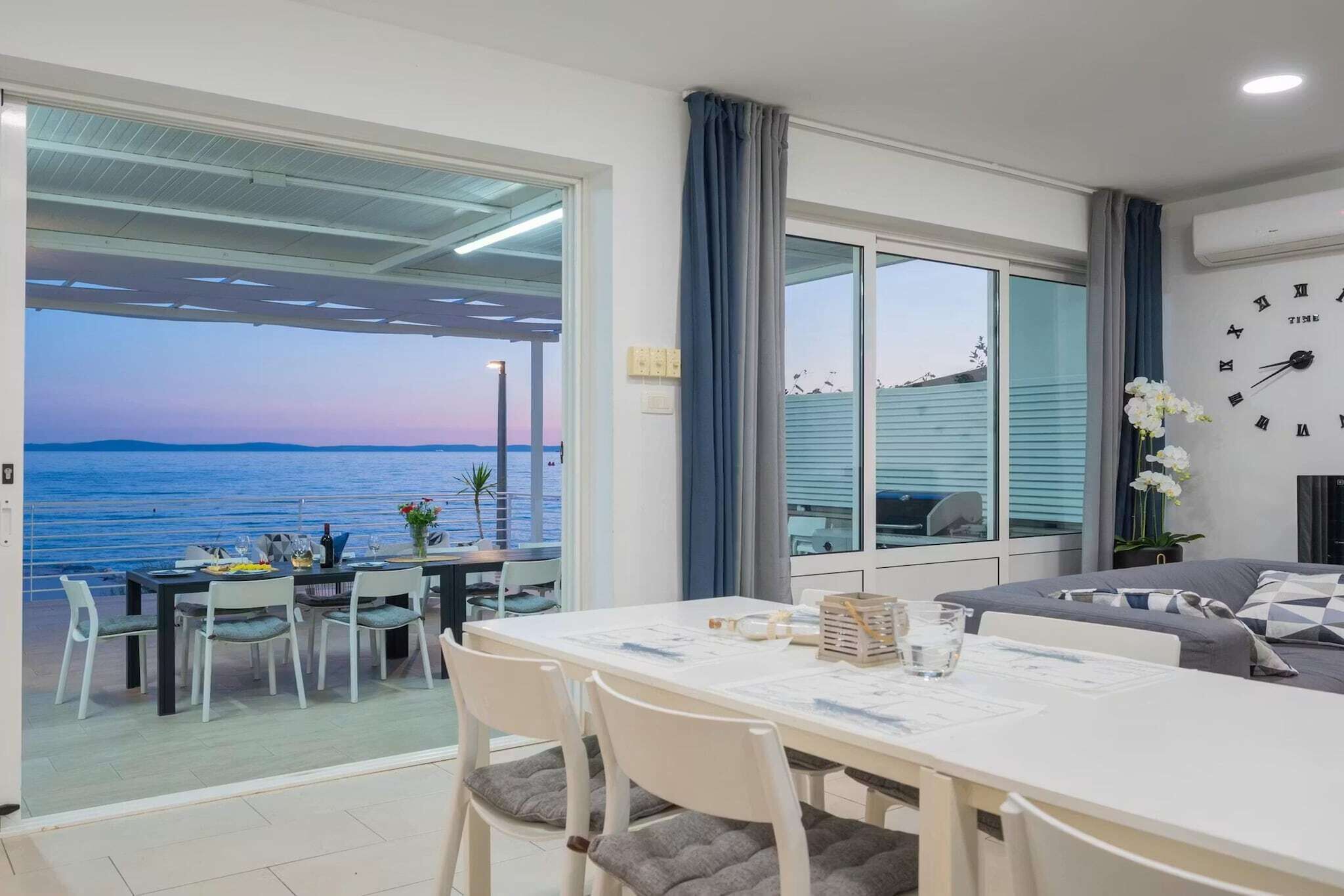 A room with a dining table, chairs, a sofa, a clock, and a view of the sea.