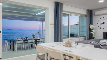 A room with a dining table, chairs, a sofa, a clock, and a view of the sea.