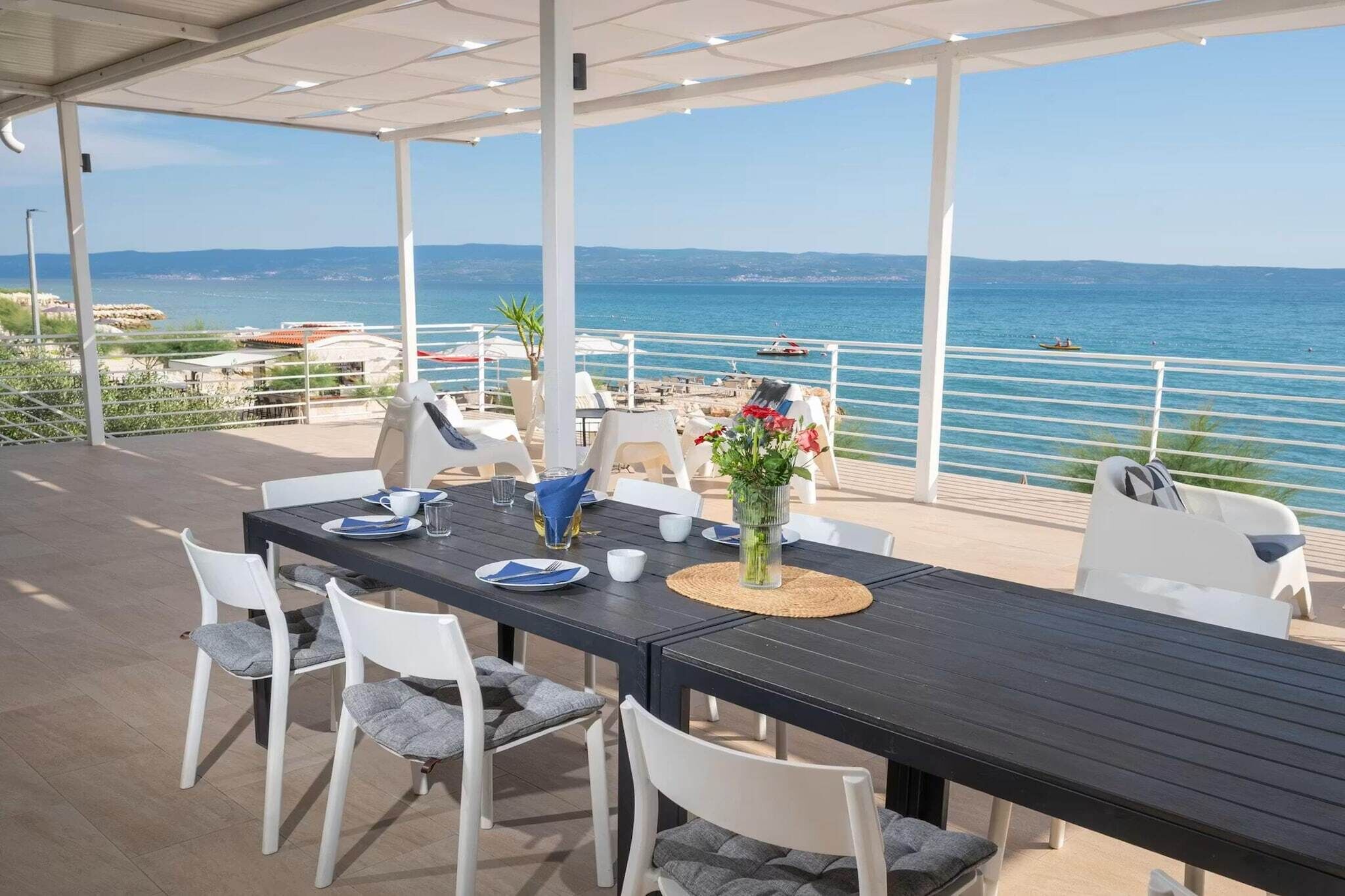 Un espace extérieur aménagé pour les repas, avec vue sur la mer, des chaises blanches, une table avec des assiettes et un vase de fleurs, ainsi qu'une terrasse couverte.
