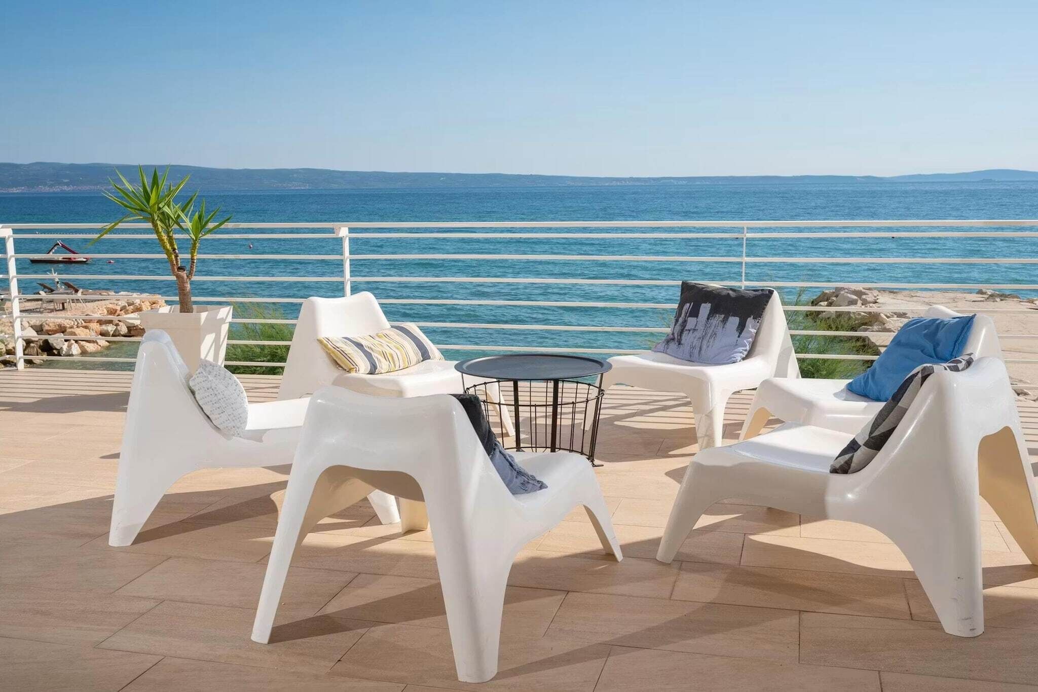 Een terras met witte plastic stoelen en een kleine ronde tafel, met uitzicht op een strand en de zee.
