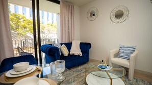 Une pièce avec un canapé bleu, un fauteuil blanc, une table basse en verre et un coin repas avec des assiettes et des verres.