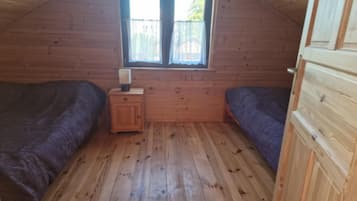 Uma cabana pequena, com um único quarto, paredes e chão de madeira, uma janela com cortinas, uma pequena mesa de cabeceira de madeira e duas camas.