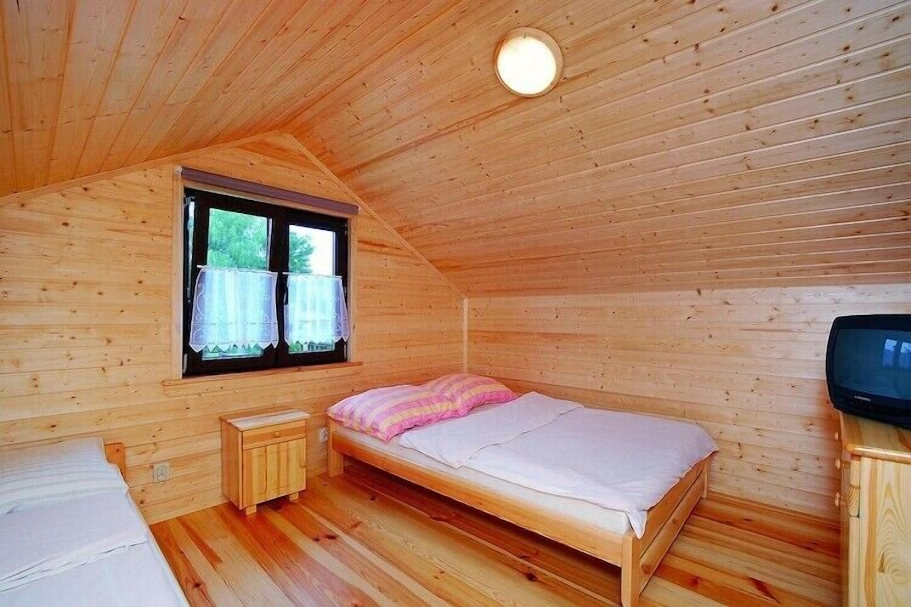 Uma cama de solteiro com um cobertor rosa, uma pequena mesa de cabeceira de madeira, uma televisão e uma janela com cortinas.