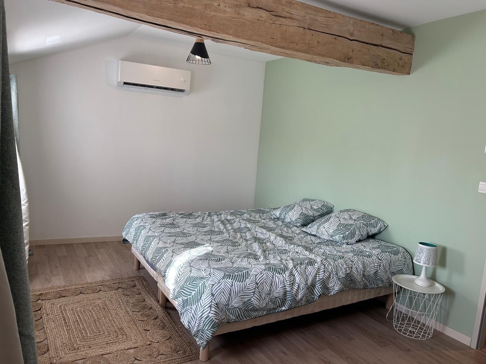 Un dormitorio con cama, aire acondicionado montado en la pared, viga de techo de madera, alfombra tejida y una mesita de noche con lámpara.