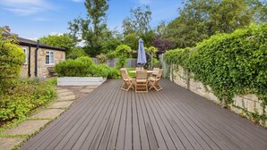 Una terraza de madera con una mesa y sillas, rodeada de follaje y un muro de piedra.