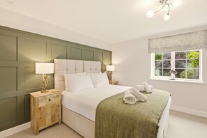 A bedroom with a large bed, two bedside tables with lamps, a window with a view of greenery, and a green herringbone-patterned bedspread.