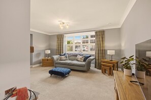 A living room with a grey sofa, blue ottoman, and a wooden coffee table. There is a window with curtains, a lamp, and a plant. - The Cottage - Powburn - North East Escapes (Powburn)