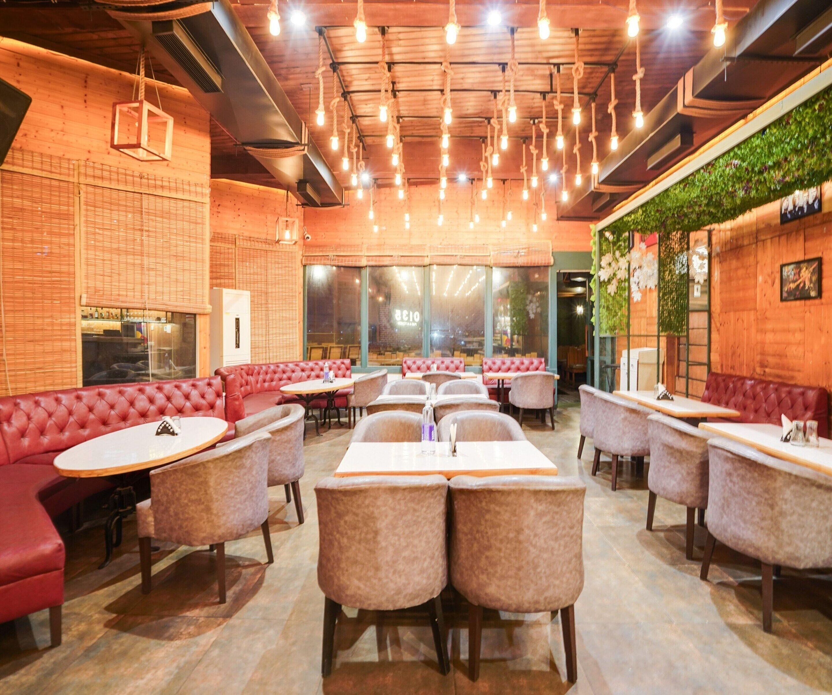 A restaurant interior with red cushioned booths, round tables, hanging light fixtures, and a green wall with plants.