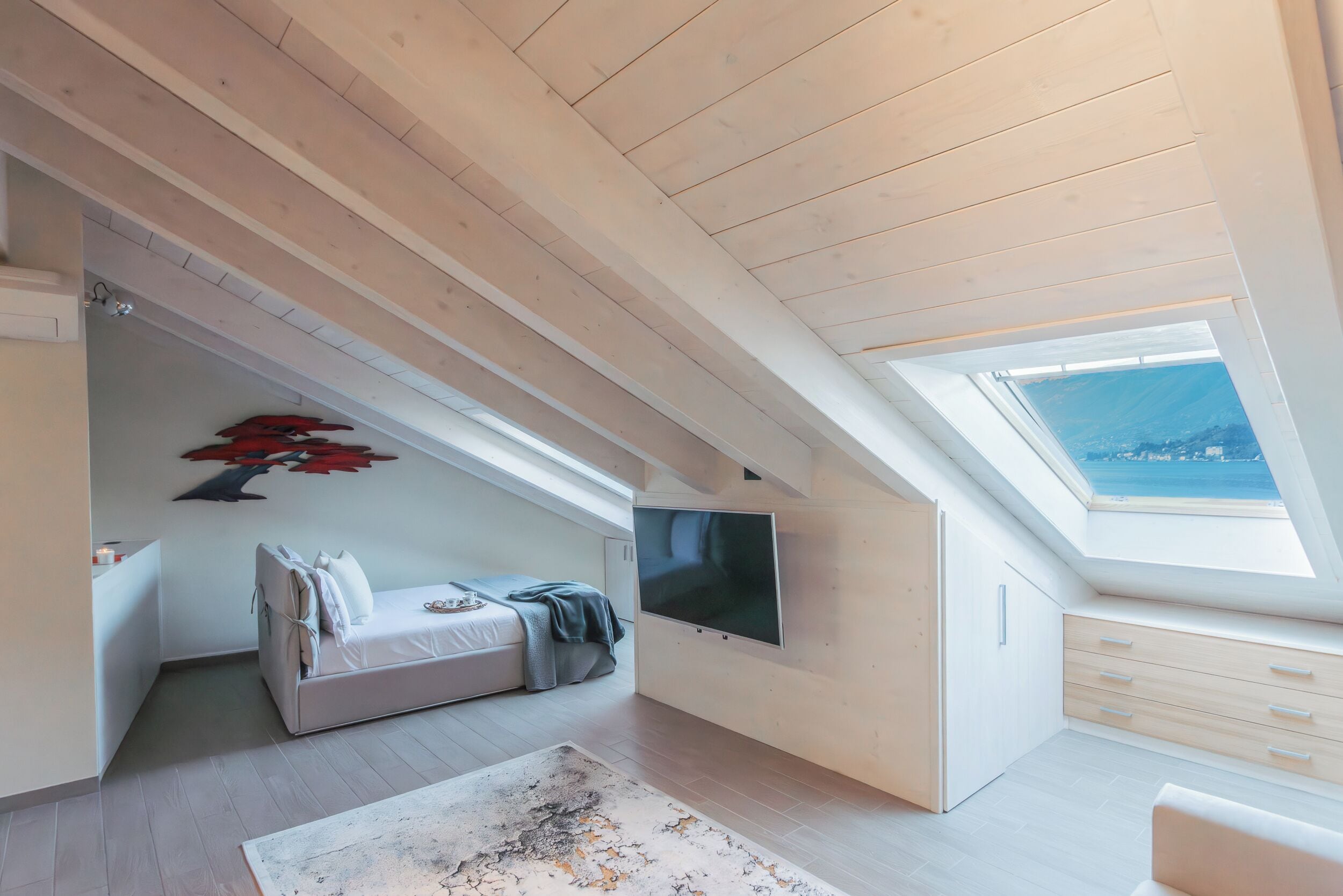 A modern loft bedroom with a sloped ceiling, a large TV, a bed, a desk, and a view through a skylight.
