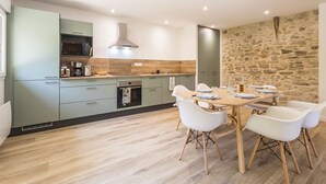 A modern kitchen with light green cabinets, a wooden countertop, and a stone wall. There is a dining table with white chairs and a wooden floor. - Peaceful country cottage - 6 guests (Nort-sur-Erdre)