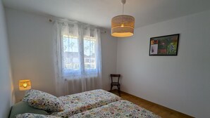 Een slaapkamer met twee bedden, een raam met gordijnen, een hanglamp, een stoel en een lijst met een afbeelding aan de muur.