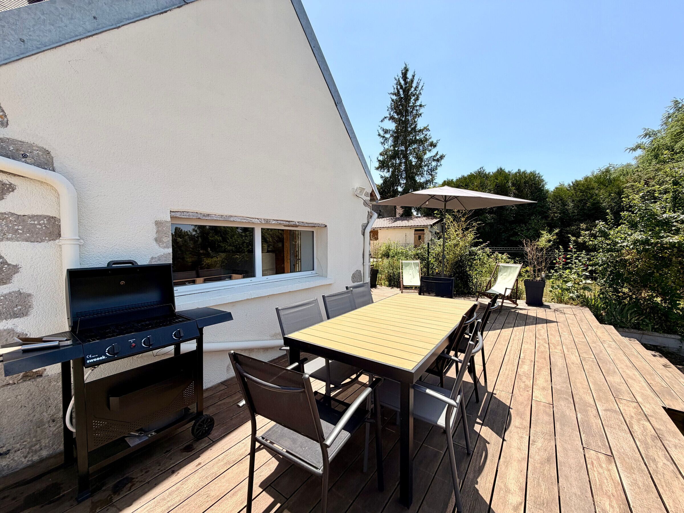 Een terras buiten met een barbecue, houten tafel, stoelen en een bloempot.