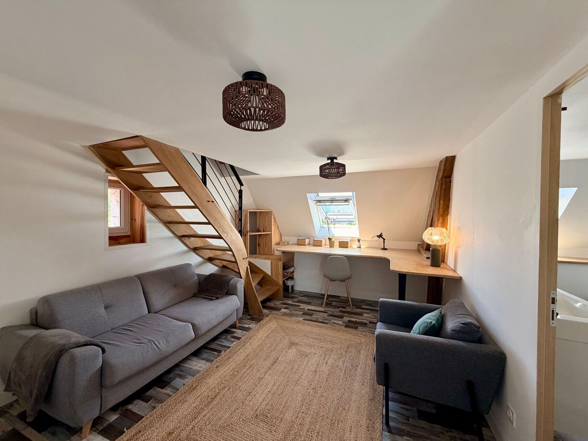A loft-style room with a wooden staircase, a gray sofa, a small desk, and a large window.