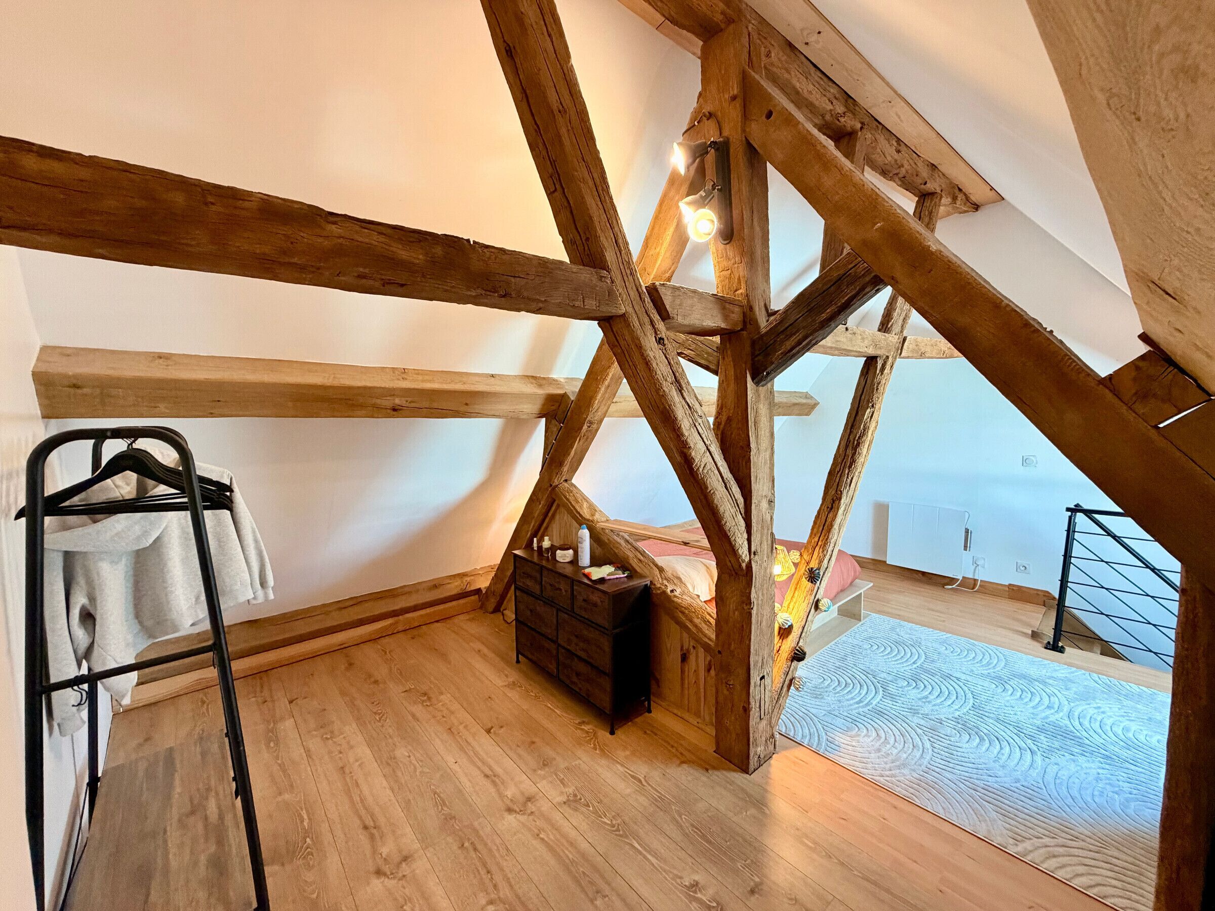 Slaapkamer in een loft met zichtbare houten balken, een hangende kledingrek, een klein houten kistje en uitzicht op de omgeving.