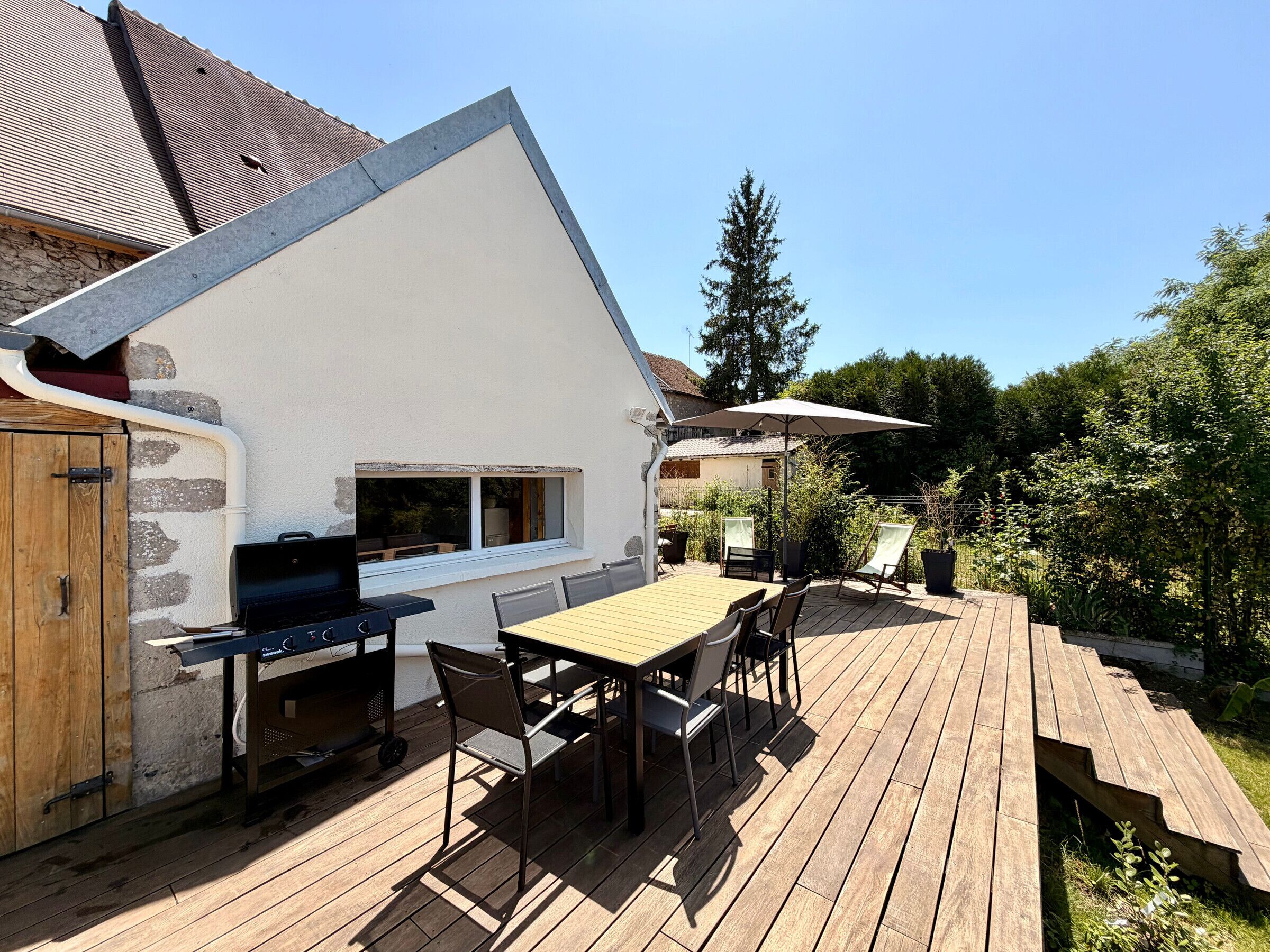Eine Terrasse im Freien mit Grill, Tisch und Stühlen, umgeben von einer Holzterrasse und einer Steinmauer.