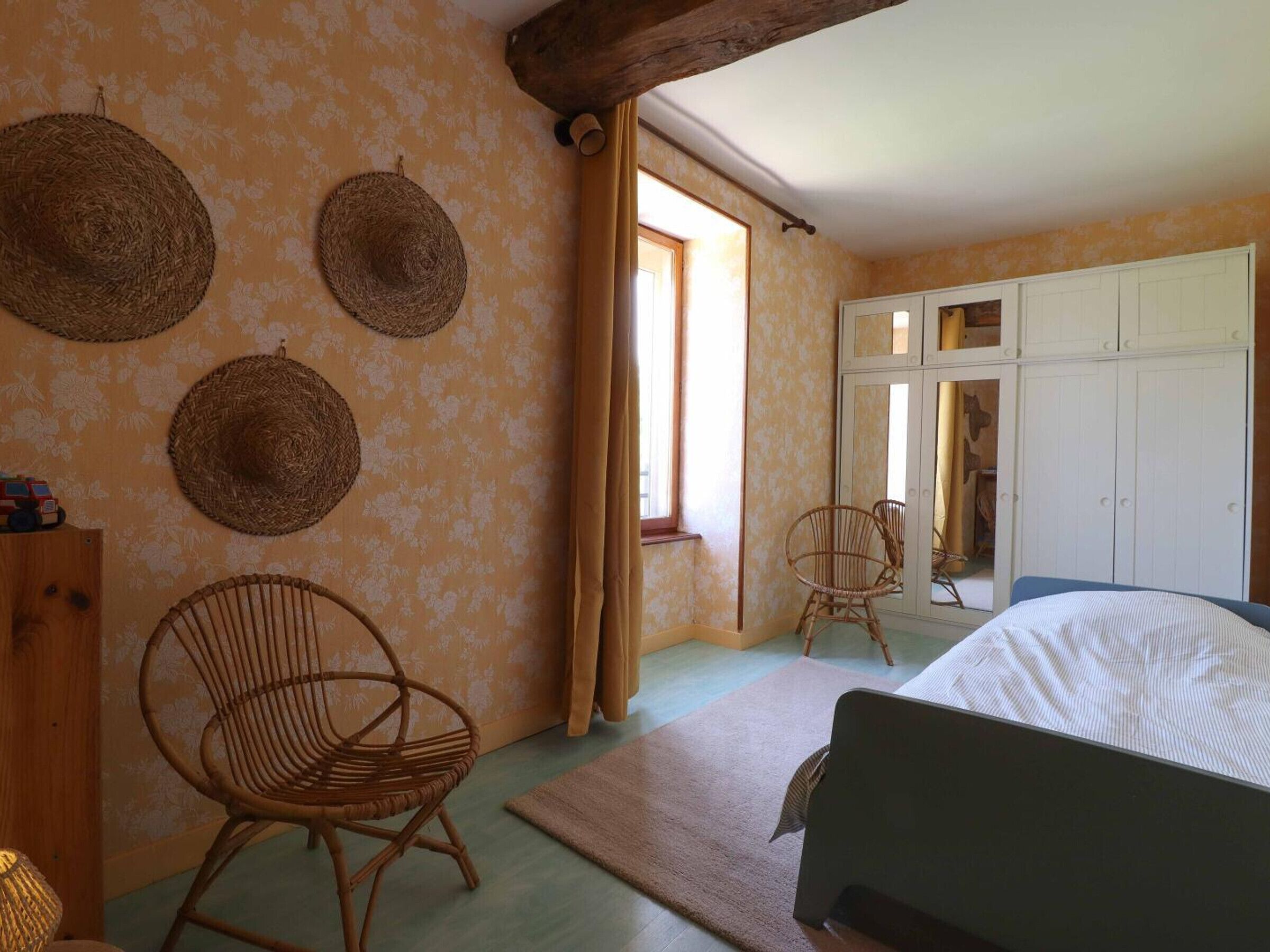 Quarto com uma cama, duas cadeiras de vime, um guarda-roupa de madeira e uma janela com cortinas.