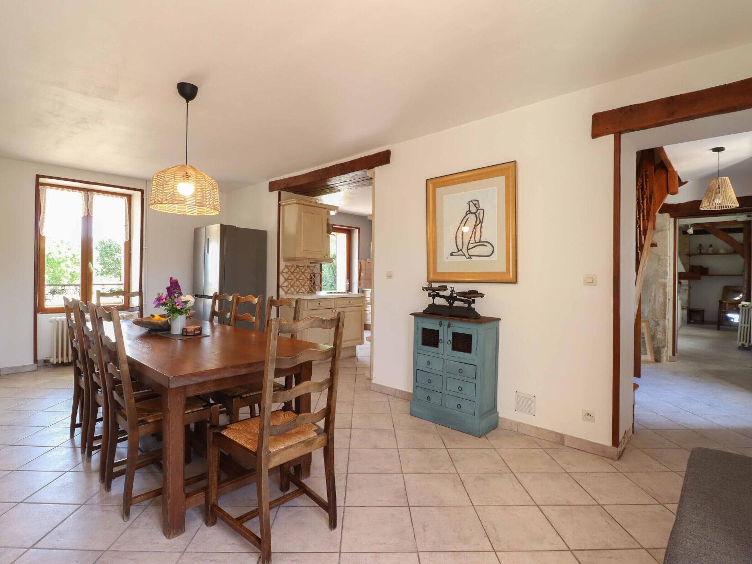 A dining area with a wooden table and chairs, a kitchen in the background, and a framed picture on the wall.