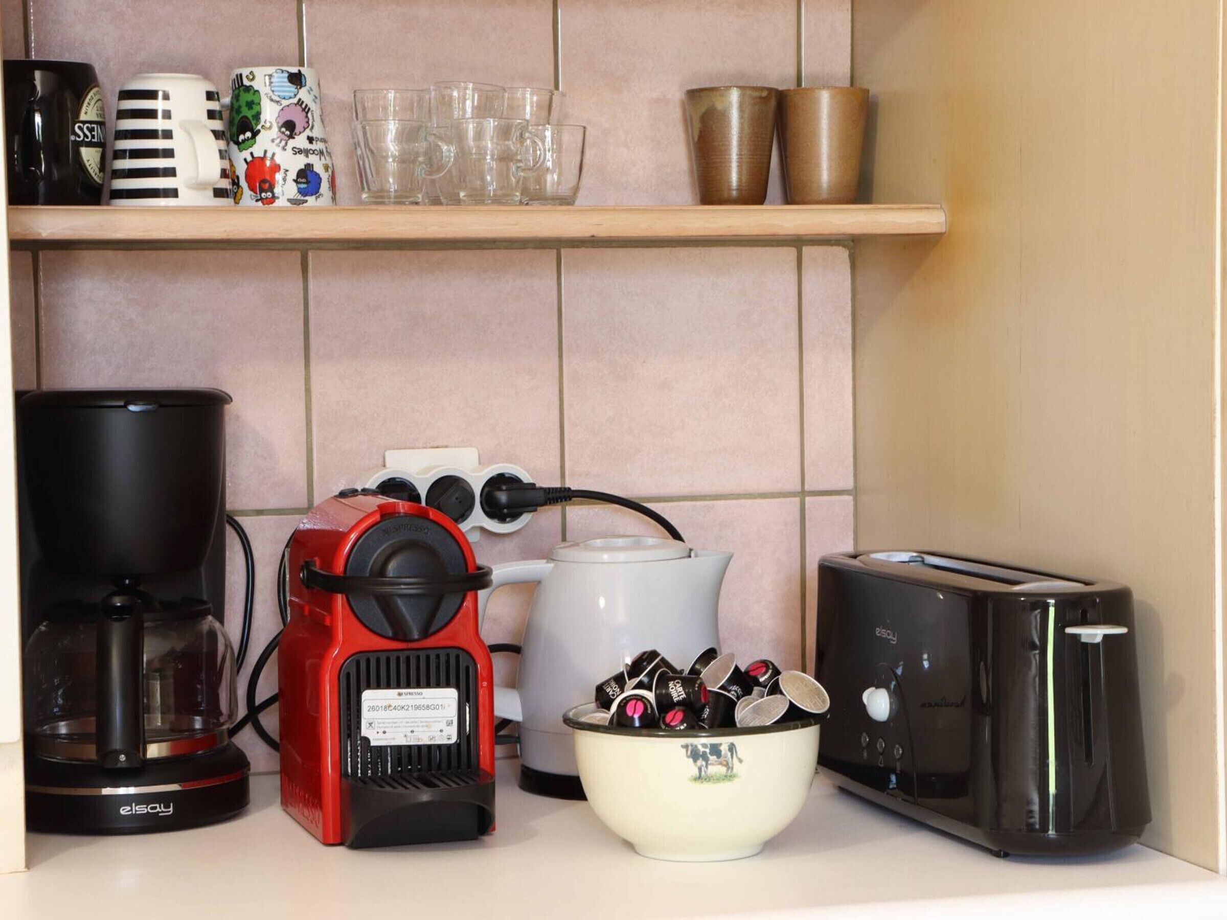 A kitchen counter with a coffee maker, a red espresso machine, a bowl of coffee pods, a toaster, and various mugs and cups on a shelf.