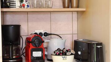 A kitchen counter with a coffee maker, a red espresso machine, a bowl of coffee pods, a toaster, and various mugs and cups on a shelf.