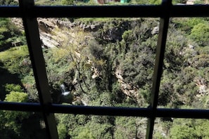 A view through a window with a metal grid, featuring a natural landscape with a waterfall, dense greenery, and rocky terrain. - Cau de lárt (Llucena)