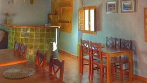A dining area with wooden tables and chairs, a kitchen with a green backsplash, and a tiled floor.