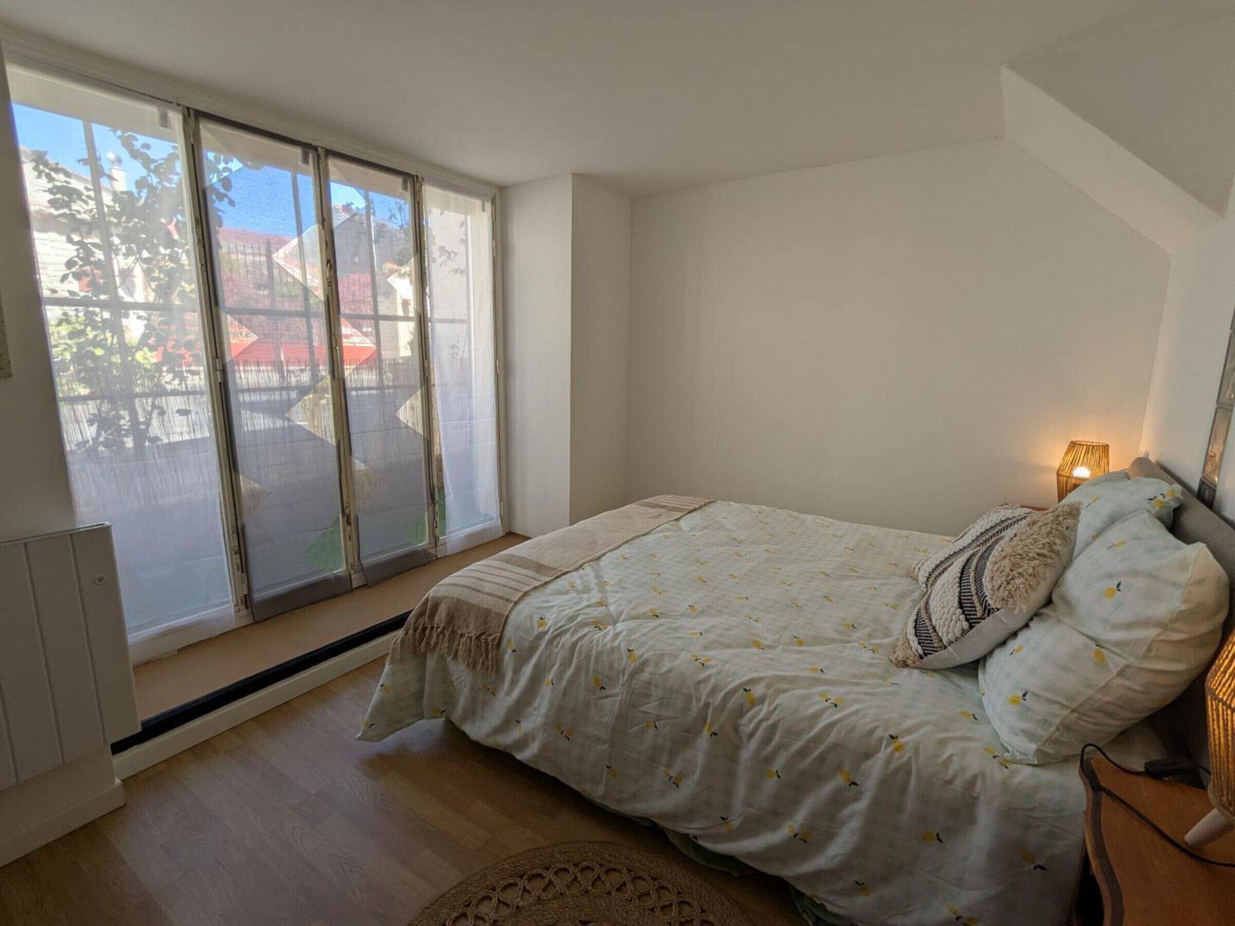 A bedroom with a large window, a bed with a patterned bedspread, a bedside lamp, and a wooden floor.