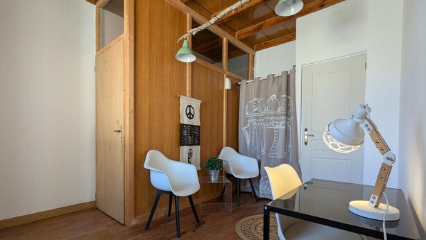 Ein Zimmer mit Holzboden, einem Tisch mit Stühlen, einem Schreibtisch mit Lampe, einer Friedenssymbol-Dekoration und einem Vorhang mit Muster.