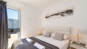 Quarto de hotel com uma cama, duas mesas de cabeceira, um quadro e vista através de uma porta de vidro.
