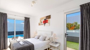 Quarto moderno com cama, mesa de cabeceira e vista para uma piscina e vegetação.
