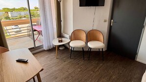 Quarto com televisão na parede, duas cadeiras de vime, uma pequena mesa e uma porta.