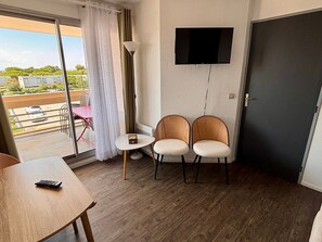 A room with a TV mounted on the wall, two wicker chairs, a small table, and a door. - Lovely air-conditioned apartment - 4 guests, mountain view (Canet-en-Roussillon)