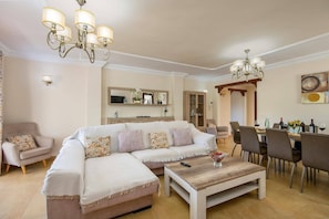 A living room with a sectional sofa, a coffee table, a dining area with chairs, and a chandelier. - Villa Lara Pinta by Villa Plus (Nerja)