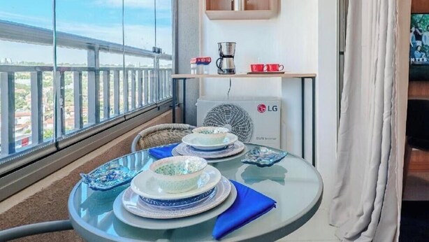 A balcony with a table set for dining, a fan, and a shelf with a basket and a kettle. - Fully furnished apartment with TV and free internet. (São Paulo)