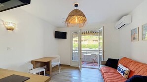A compact room with a wooden table, a white chair, a sofa with patterned cushions, a hanging woven light fixture, and a television on the wall. - Studio with AC and terrace - 4 guests (Saint-Cyr-sur-Mer)