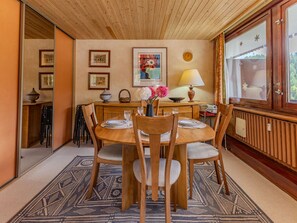 A dining room with a wooden table, chairs, a patterned rug, framed pictures, and a window with curtains. - Cozy apartment, mountain view - 6 guests. (La Plagne)