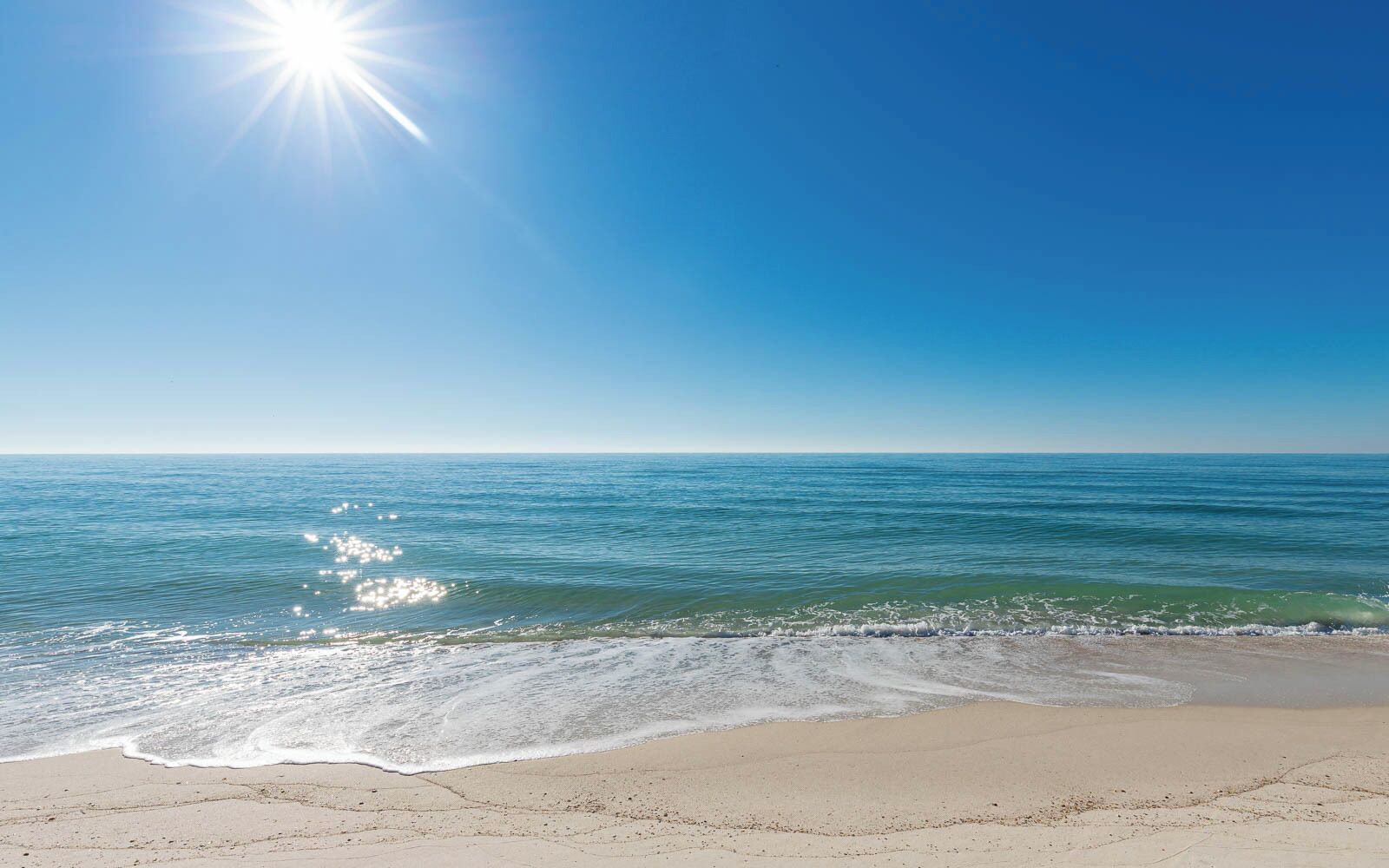 Ein Strand mit blauem Himmel und Sonnenlicht, das sich im Wasser spiegelt.