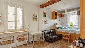 Een kleine kamer met een bureau, twee stoelen, een bank, een bed en een kitchenette.