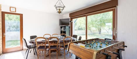 Un comedor con mesa de fútbol, sillas, televisor y ventana con vista a follaje.