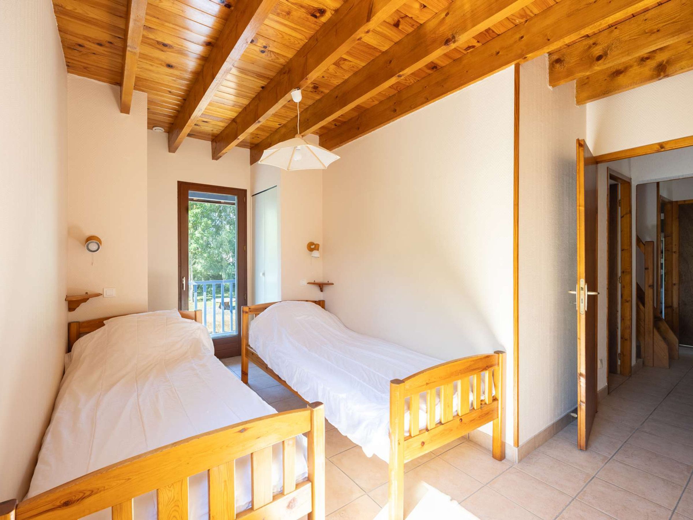 Een kamer met twee houten bedden, een plafond met zichtbare balken, een deur die naar buiten leidt, en een klein plankje met een lamp.