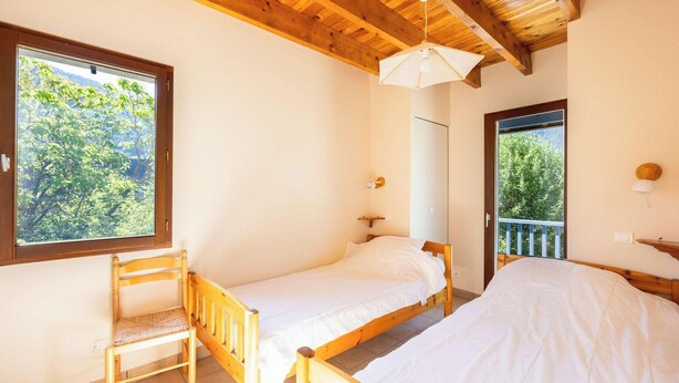 Une chambre avec deux lits simples, une chaise en bois, une fenêtre donnant sur des arbres et un ventilateur de plafond.