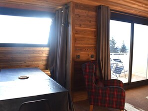 A wooden cabin interior with a table and plaid chairs, a window with curtains, and a view of the outdoors. - Apartment panoramic Pyrenees view - guests (Font-Romeu-Odeillo-Via)